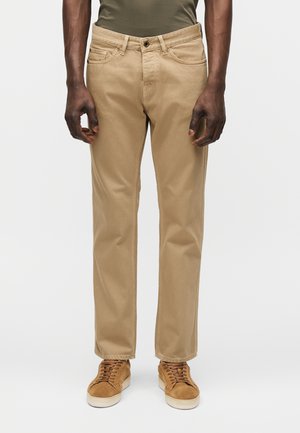 Mężczyzna w beżowych spodniach o prostych nogawkach, oliwkowej włożonej do środka koszulce i jasnobrązowych zamszowych trampkach z białą podeszwą.