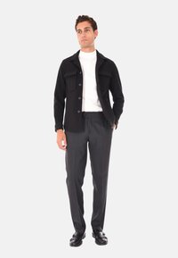 Giacca nera con bottoni, due tasche frontali, dolcevita bianco, pantaloni grigi e scarpe nere. Modello in piedi davanti a uno sfondo grigio chiaro.