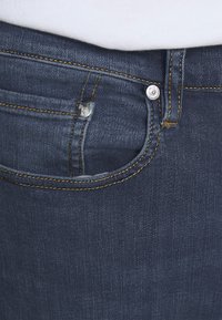 Mörkblå denimjeans på nära håll som visar framfickan, bältesöglan och metallknappen med zebra-gravering, bärs med en vit skjorta.