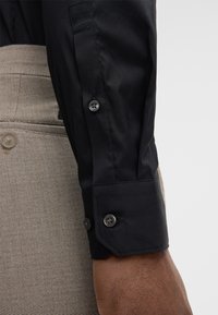 Manica di una camicia nera con polsino abbottonato e pantaloni beige testurizzati visibili sullo sfondo, sul braccio di una persona.
