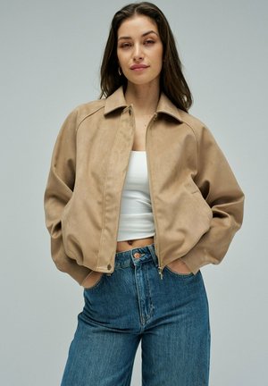 Femme portant une veste aviateur en daim beige, un débardeur blanc court et un jean bleu taille haute, debout les mains dans les poches de la veste.