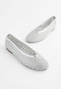 Ballerines argentées avec clous ronds brillants, petit talon et doublure intérieure à pois gris sur fond blanc.