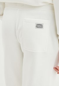 Witte sweatpants gemaakt van zacht materiaal, met een achterzak en een zwart etiket met de tekst "CREEKS," waarmee het merk wordt benadrukt.