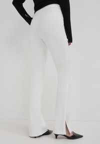 Persona che indossa pantaloni bianchi a gamba dritta con aperture laterali all'orlo, scarpe nere con tacco a stiletto e una maglietta nera a maniche lunghe.