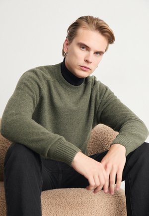 Olive green ribbed sweater with a high neckline, worn over a black turtleneck, paired with dark jeans, seated on a textured beige chair.