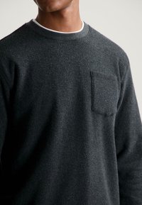 Uomo che indossa un maglione grigio scuro a girocollo con tasca sul petto sopra una maglietta bianca, mostrato dalla zona del collo fino a metà torso su uno sfondo semplice.