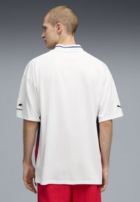 Άνδρας με την πλάτη γυρισμένη φοράει λευκό πουκάμισο Puma Motorsports με λογότυπα στα μανίκια και κόκκινα σορτς, μπροστά σε γκρι βαθμιδωτό φόντο.