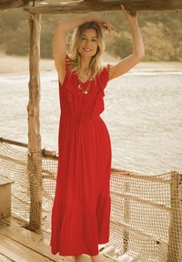 Red maxi dress with ruffled sleeves, cinched waist, and a tiered skirt. Model stands against a coastal backdrop, with natural textures and colors.