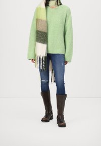 Pull vert texturé surdimensionné, jean skinny bleu déchiré, bottes marron montant jusqu'aux genoux, et écharpe multicolore à motifs avec franges.