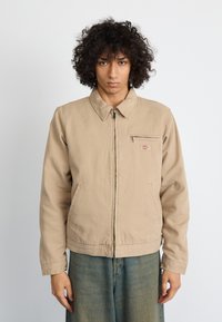 Beige jacket with a zip front and pointed collar, featuring a chest pocket and side pockets, made of durable fabric with a smooth texture.