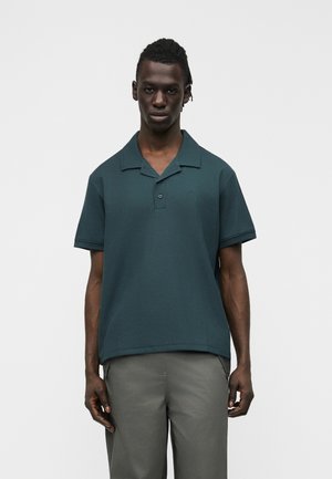 Άνδρας που στέκεται μπροστά σε απλό φόντο, φοράει κοντομάνικο πόλο σε σκούρο πράσινο χρώμα και γκρι παντελόνι, κοιτώντας μπροστά με ουδέτερα έκφραση.