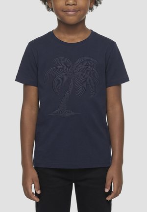 Enfant portant un t-shirt bleu marine avec un motif de palmier en relief, debout devant un fond clair uni.