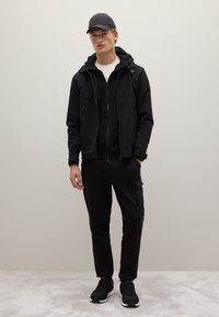 Schwarzes Sportoutfit mit einer hoffeingezippten Jacke, die über einem schwarzen Shirt getragen wird, kombiniert mit Jogginghosen und schwarzen Sneakers.