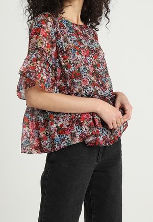 Mujer vistiendo una blusa floral de manga corta y suelta con mangas con volantes, combinada con pantalones negros de cintura alta sobre un fondo claro.