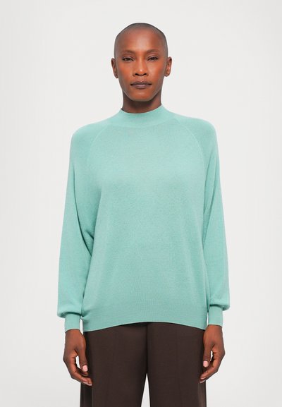 Pull en maille légère vert menthe, col haut, manches raglan, poignets et ourlet côtelés avec une coupe décontractée, associé à un pantalon marron foncé.