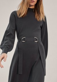 Langdurige, donkergrijze jurk met lange mouwen en een hoge hals, getailleerde taille met zilveren accessoires en vloeiende stof. Soepele textuur met gedrapeerde details.