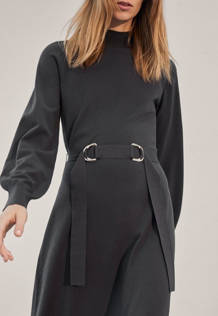 Langdurige, donkergrijze jurk met lange mouwen en een hoge hals, getailleerde taille met zilveren accessoires en vloeiende stof. Soepele textuur met gedrapeerde details.