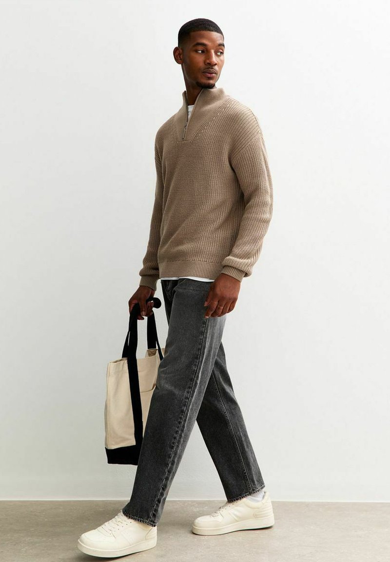Beige gebreide trui met een halve rits kraag, gecombineerd met grijze jeans en witte sneakers. Houdt een beige schoudertas met zwarte banden vast.