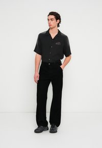 Μαύρο κοντομάνικο πουκάμισο με κουμπιά στο μπροστινό μέρος και διακριτικό λογότυπο στο στήθος, συνδυασμένο με μαύρο φαρδύ παντελόνι από κορδόνι και σκούρα αθλητικά παπούτσια.