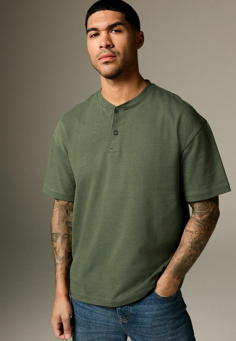 Short-sleeve olive green henley shirt with two-button placket, made of soft fabric, paired with blue denim jeans.