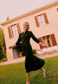 Γυναίκα με μακρύ μαύρο φόρεμα, ξυπόλυτη, χαμογελαστή και περπατάει στο γρασίδι μπροστά από ένα σπίτι με πέτρινα τοιχώματα, ξύλινα παντζούρια και υπαίθρια έπιπλα.