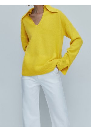 Personne portant un pull jaune vif à col en V large et un pantalon blanc, debout avec une main dans la poche sur un fond uni.