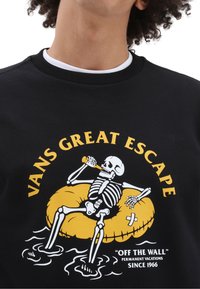 Sudadera negra con un esqueleto relajándose en un flotador amarillo, sosteniendo una bebida. El texto dice "VANS GRAN ESCAPADA" encima de la gráfica.