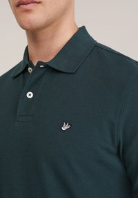Man draagt een donkergroen poloshirt met witte knopen en een klein geborduurd vogel-logo op de borst, gefotografeerd tegen een neutrale achtergrond.