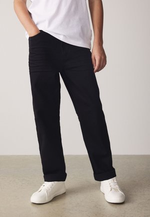 Person, der schwarze Hosen mit aufgekrempelten Säumen, weiße Sneakers und ein weißes Hemd trägt, steht mit einer Hand in der Tasche auf einem neutralen Boden.