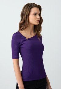 Top viola a costine con scollo a barca, maniche corte e un accent decorativo in metallo sulla spalla. Tessuto liscio, design aderente.
