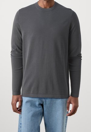 Mand iført en mørkegrå langærmet bluse med rund hals og lyseblå denimjeans, stående foran en ensfarvet baggrund.