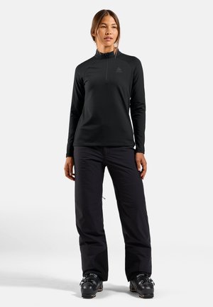 Femme debout portant un haut noir à manches longues zippé, un pantalon noir et des chaussures de ski sur un fond blanc uni.