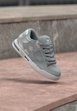 Adidaș sport gri cu accente argintii în formă de stelă și șireturi, prezentat plutind deasupra unei suprafețe de piatră lângă un zid de beton.