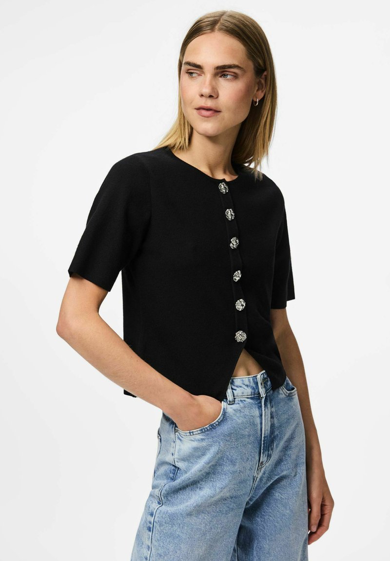 Melnas īsām piedurknēm kardigāns ar apaļu izgriezumu, kam pa priekšā ir sudraba ziedu pogas, apvienots ar gaiši zilo augsto jostasvietu džinsu.