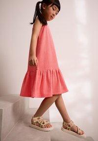 Robe corail texturée avec un ourlet évasé, associée à des sandales à brides multicolores ornées de broderies. Enfant marchant sur des escaliers.
