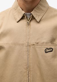 Chaqueta de algodón beige con cierre de cremallera, cuello puntiagudo y parche de logo negro en el lado izquierdo. Costuras reforzadas a lo largo de las costuras.