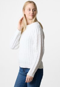 White cable-knit sweater with long sleeves and ribbed hem. Worn over blue jeans, showcasing textured pattern and fitted design.
