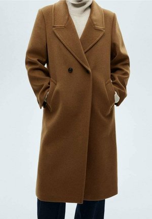 Manteau brun en laine à double boutonnage avec de larges revers, porté sur un pull à col roulé blanc et un pantalon foncé, mains dans les poches du manteau.