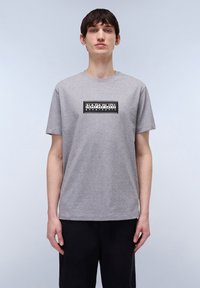 Camiseta gris de manga corta con cuello redondo. Presenta un logo rectangular negro en el pecho, hecho de una tela suave similar al algodón.