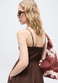 Μια ξανθιά γυναίκα με γυαλιά ηλίου και καφέ αμάνικο φόρεμα κρατά μια μεγάλη καφέ και λευκή τσάντα ώμου με σχέδια.