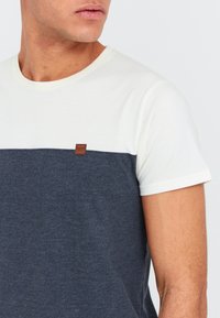Kortærmet t-shirt med hvid overdel og marineblå nederste sektion, med et lille brunt lædermærke på brystet.