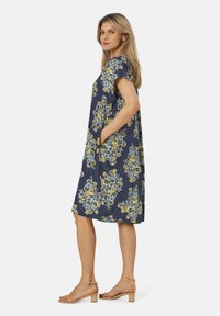 Robe florale marine à manches courtes, dotée de poches et d'une coupe décontractée ; décorée de fleurs jaunes et bleues ; associée à des sandales beiges.