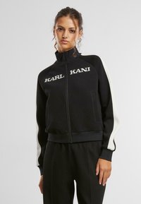 Černá zipová bunda s bílými bočními pruhy. Má vysoký límec a logo "KARL KANI" na přední straně. Hladký materiál a přiléhavý střih.
