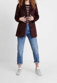 Manteau en laine bordeaux avec un col cranté et deux poches avant, porté sur une chemise rayée, associé à un jean bleu clair retroussé et des baskets blanches.