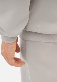 Sivý bavlnený sweatshir a tepláky s detailmi ribbed manžetami. Malá biela výšivka loga „U“ na oboch kusech. Hladký povrch.