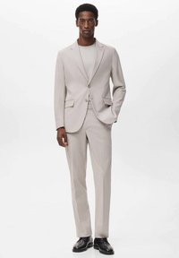 Traje gris claro con un diseño ajustado, que presenta dos botones, solapas de muesca y pantalones de frente plano. Llevado sobre una camisa clara y combinado con zapatos negros.