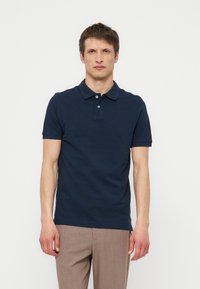 Tânăr cu păr maro scurt, poartă un tricou polo bleumarin cu mânecă scurtă și pantaloni maro deschis, stând în fața unui fundal alb simplu.