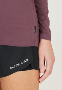 Langärmliges weinrot sportliches Oberteil mit flachen Nähten. Kombiniert mit schwarzen Shorts mit weißem "ELITE LAB"-Logo. Weiches, dehnbares Material.