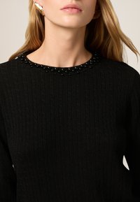 Zwarte gebreide trui met een geribbelde textuur en een ronde halslijn versierd met zwarte kralen. Het haar is losgestyled, waarbij een gouden oorbel zichtbaar is.
