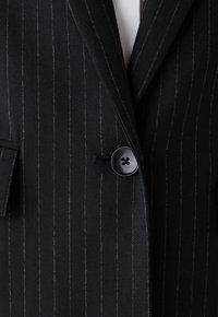 Blazer negro a rayas con un diseño estructurado, que presenta un solo botón negro y una tela texturizada. Acentos notables de costura blanca.